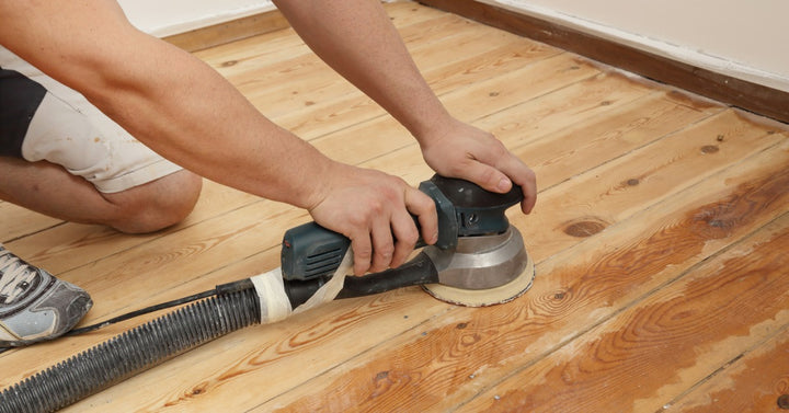 A man kneels on the floor and uses an electric sanding tool to refinish hardwood flooring during a renovation project.