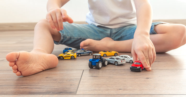 A little boy with bare feet wearing shorts and a t-shirt sits on the hardwood floor at home and plays with small toy cars.