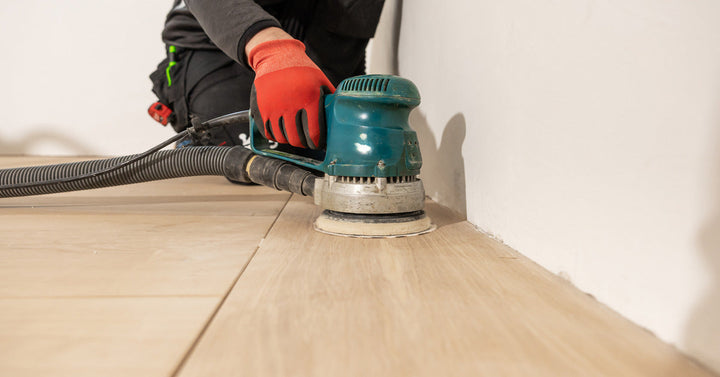 A person dressed in black clothes wears safety gloves while using a handheld sander to remove the finish from a wood floor.