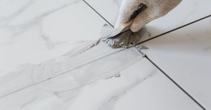 A gloved hand presses gray grout into the joints between four marbled white tiles using an off-white rubber float.