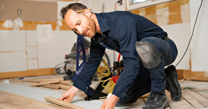 A male construction worker kneels as he installs wood-flooring panels inside a house that's being renovated.